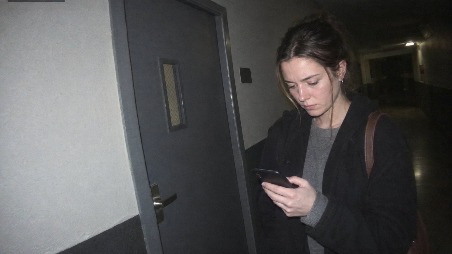 A young woman at an evening hiring event stands near a closed interview-room door, reading her phone with a serious focused expression