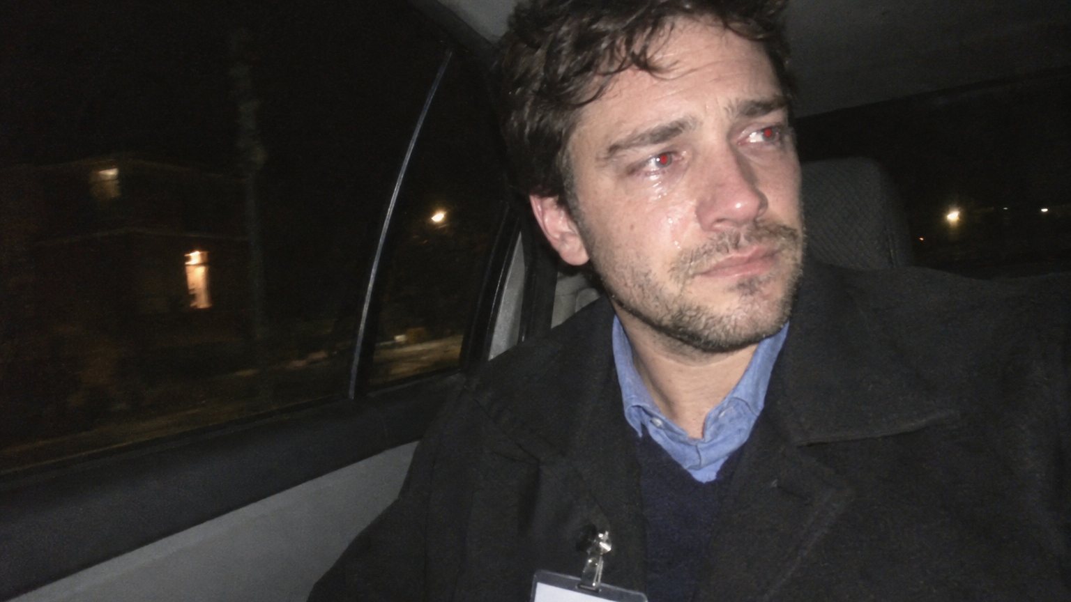 A man stands alone in a dim kitchen at night, still wearing an office-party name tag, looking away with tears welling after an awkward coworker conversation