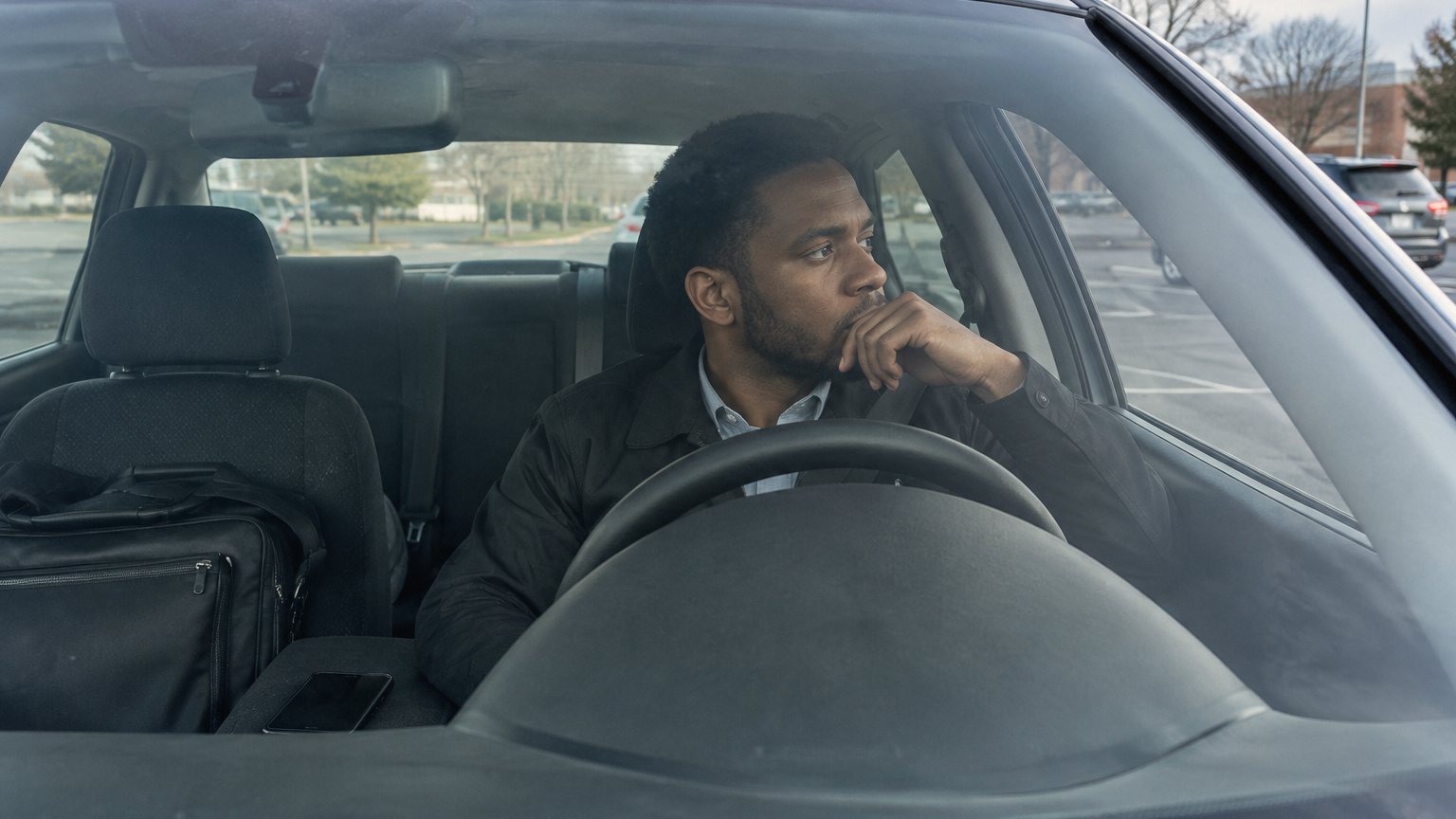A job seeker sits in a parked car after a video interview, holding back tears with the laptop bag beside him