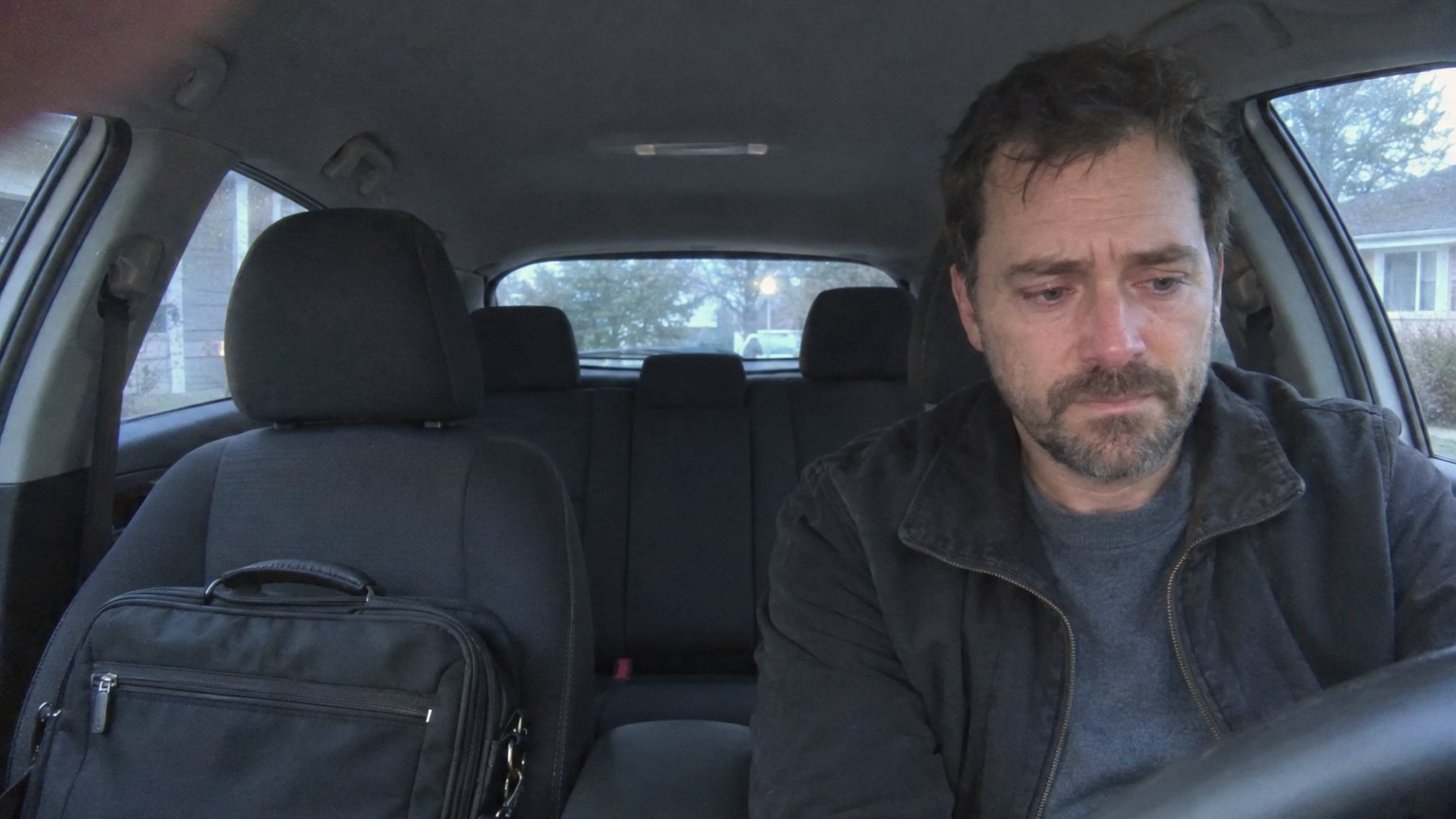 A white man sits alone in a parked car after an interview, holding back tears with a laptop bag on the passenger seat
