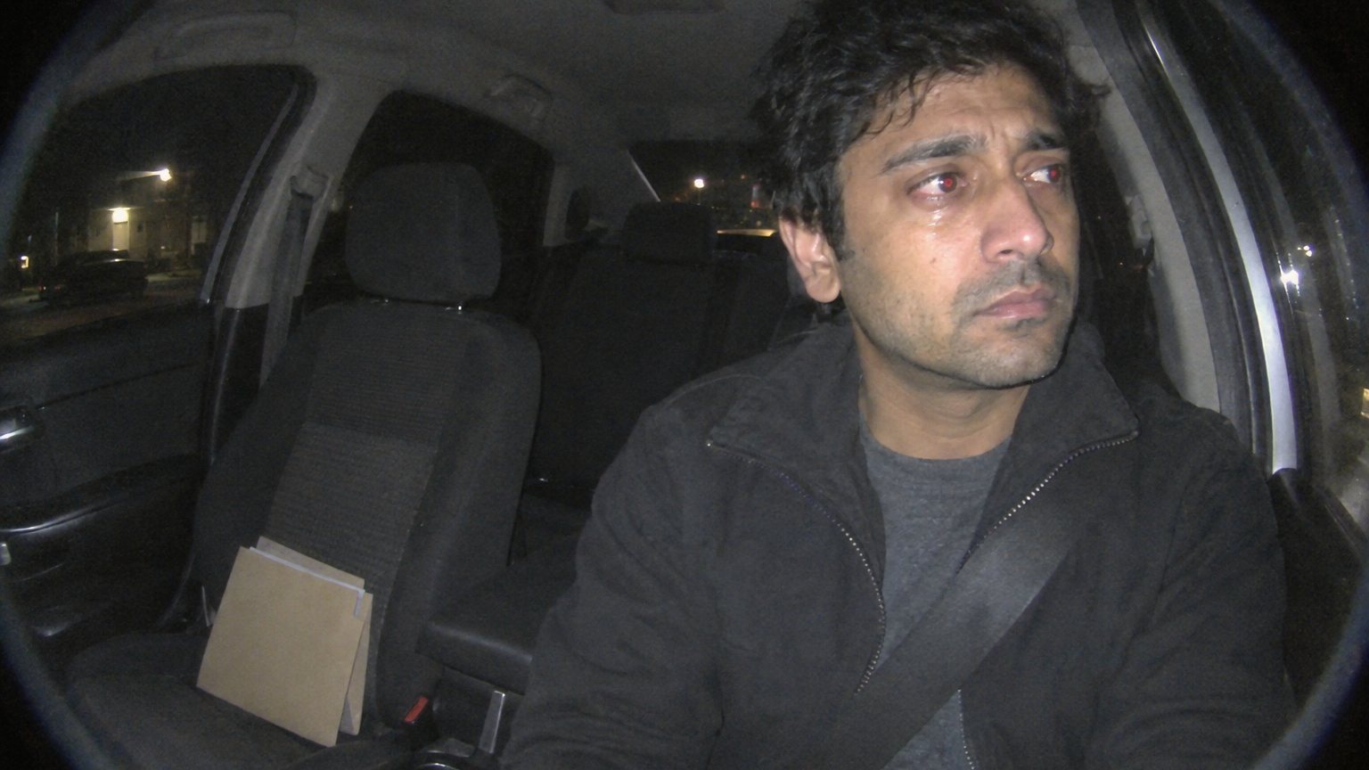 A worried South Asian man sits in a parked car at night after an interview with an older parent’s folder on the passenger seat