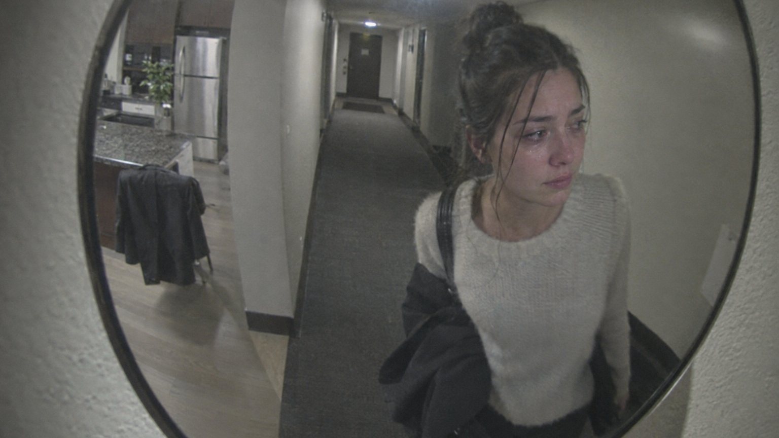 A young woman stands alone in a dark kitchen after an interview, holding back tears beside an abandoned blazer