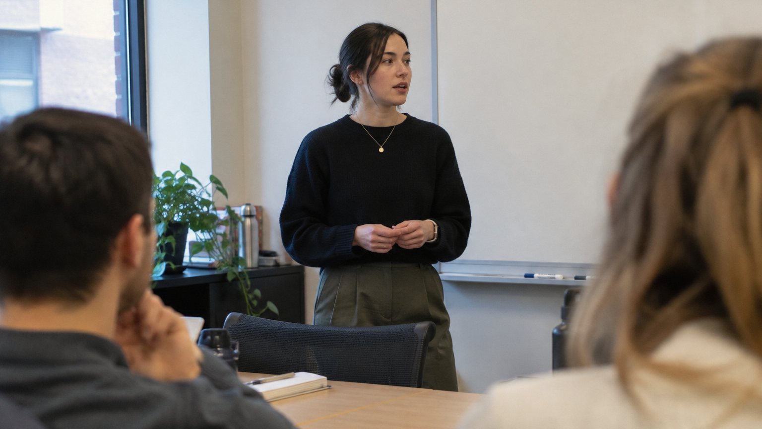 A professional stands at the front of a small meeting room while coworkers wait for her to start speaking