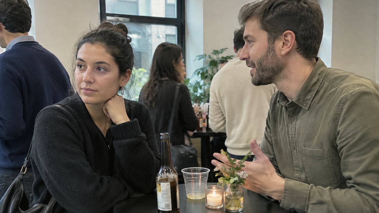 Two coworkers stand near a high-top table at an office holiday party while one briefly looks aside during conversation