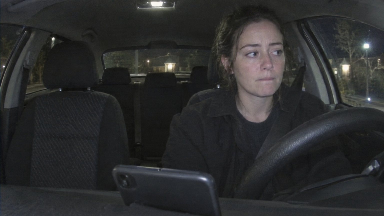 A woman sits alone in her car at night after an important interview, trying to stay calm with her phone propped on the dashboard