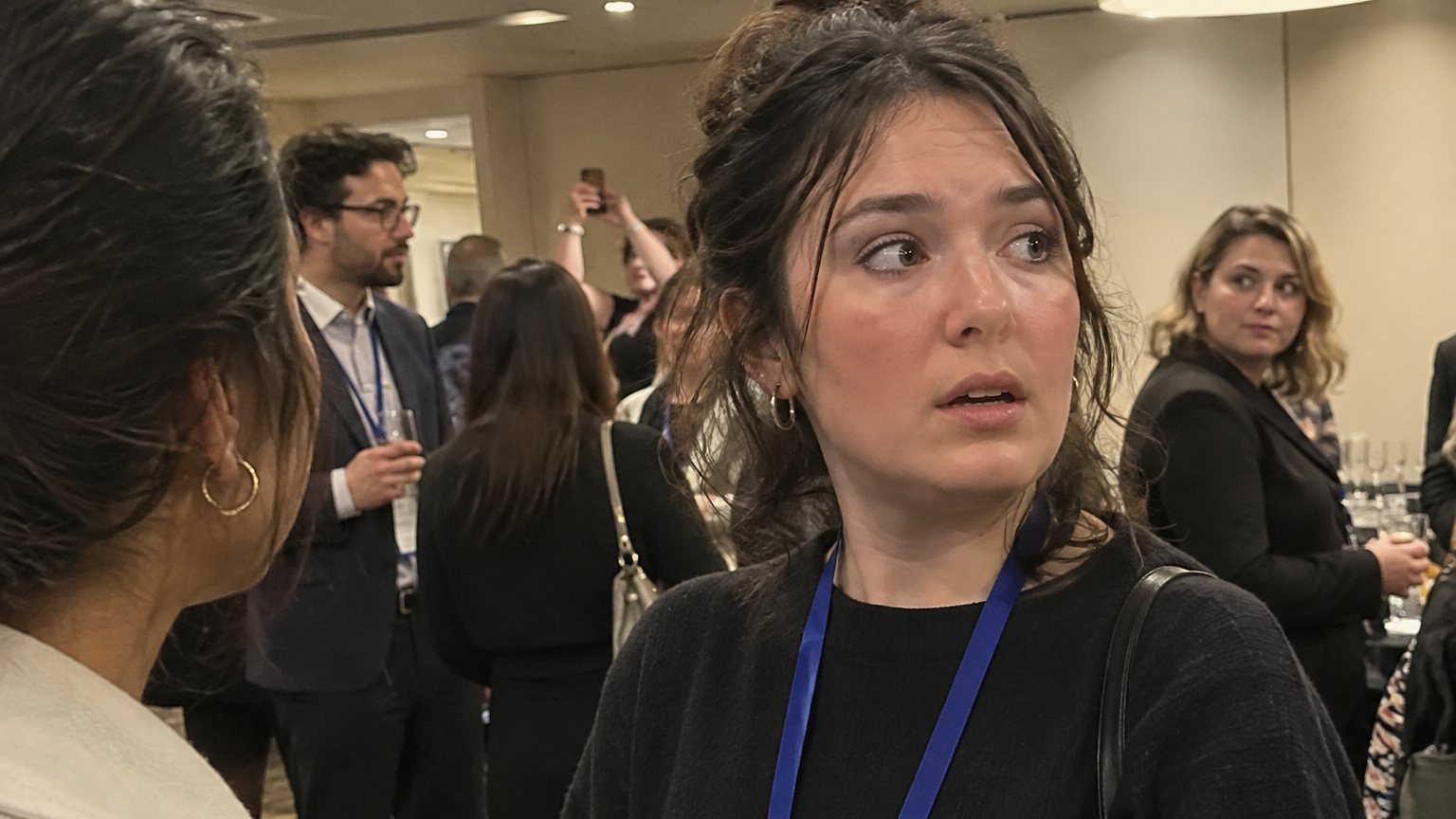 A professional at a networking reception pauses mid-conversation while a phone is raised in the background