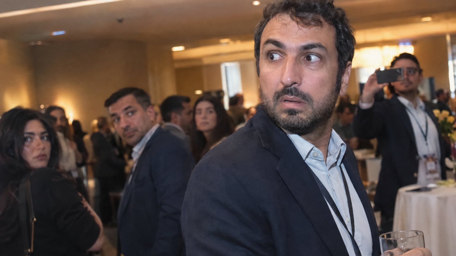 A Middle Eastern man holding a drink at a networking event looks guarded as someone across the room raises a phone
