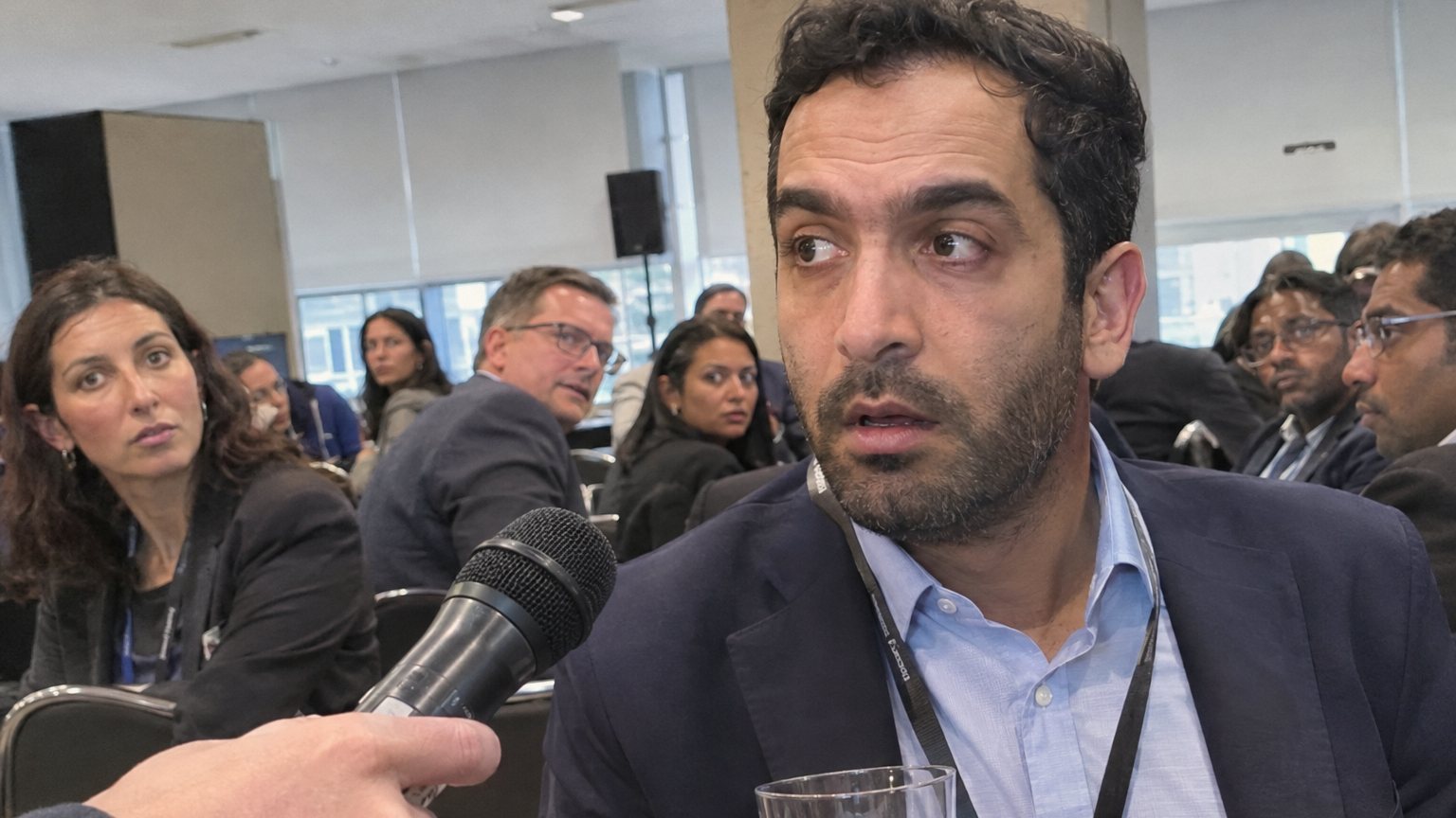 A Middle Eastern man freezes while holding a drink at a networking event as other attendees turn toward him and a microphone is offered