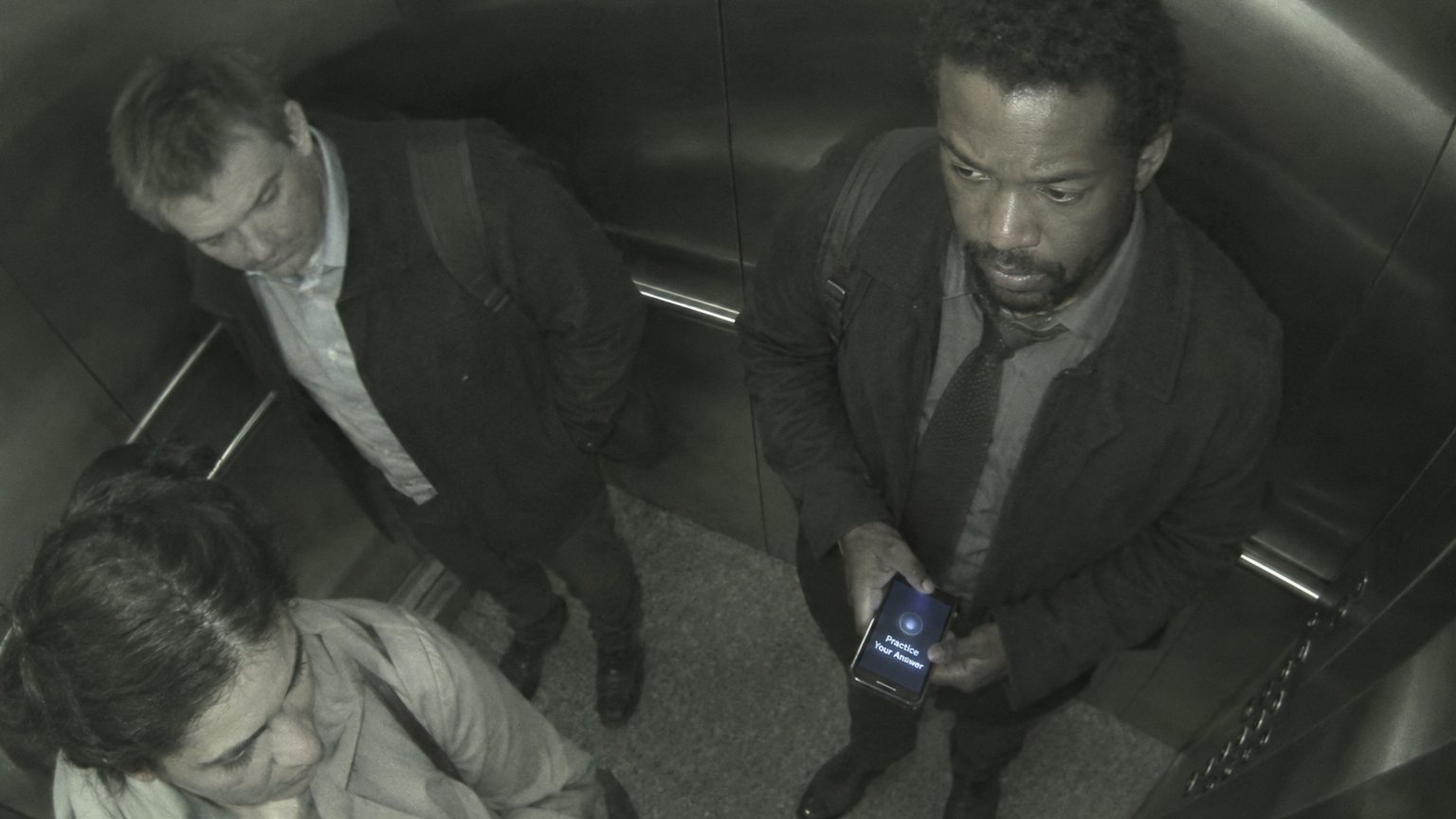 An elevator security camera view shows a tense Black man in interview clothes rehearsing quietly while holding a phone in a crowded lift