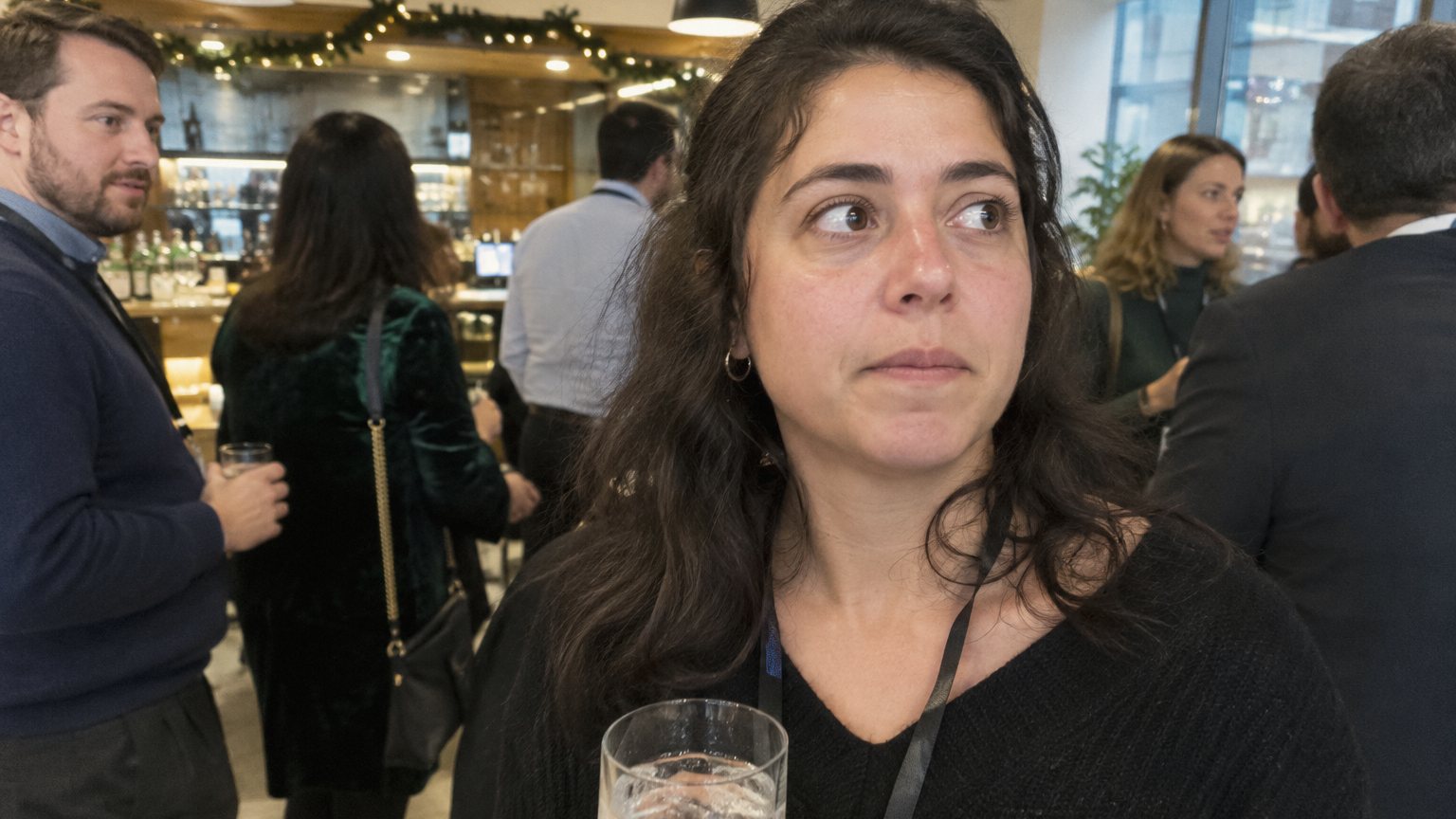 A professional at a crowded office holiday party holds a glass of seltzer near a bar while coworkers talk in the background
