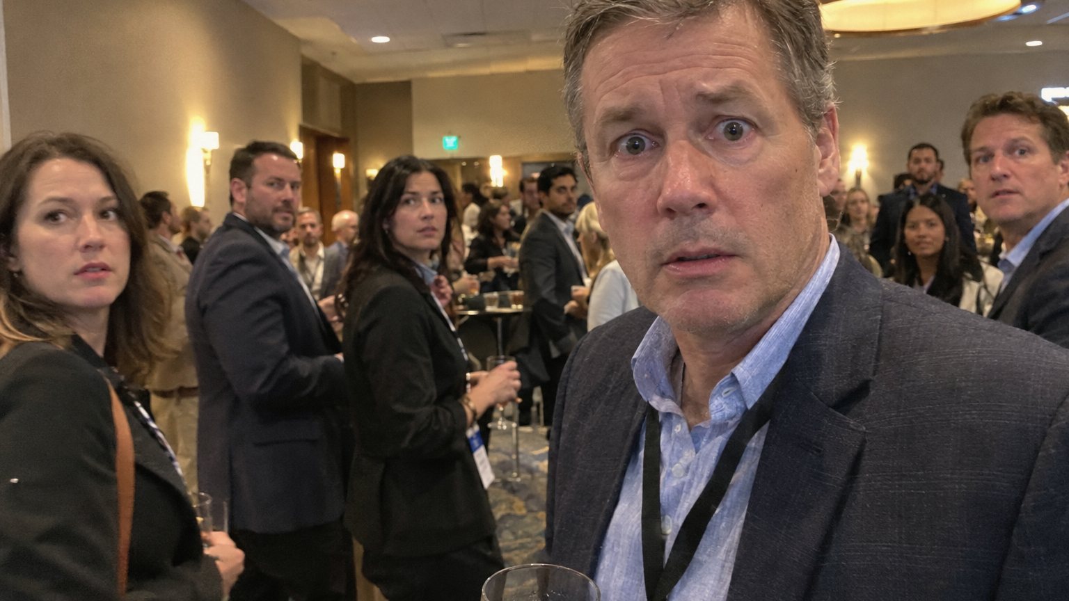 A man at a networking event holds a drink and makes uneasy eye contact while nearby attendees wait in the blurred background