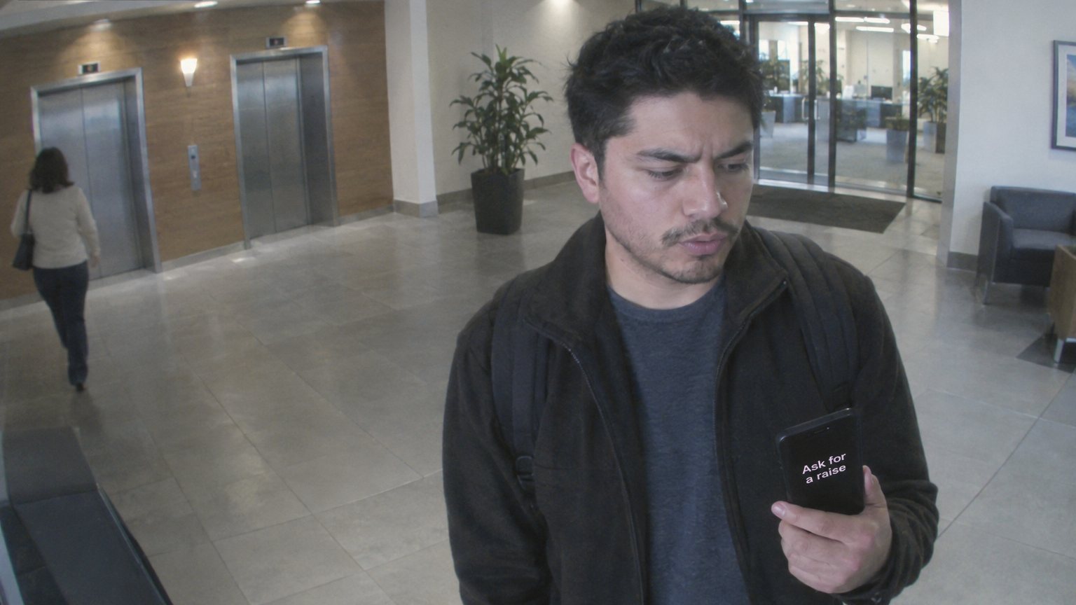 A Latino office worker sits on stairwell steps rehearsing before a pay conversation, seen from a security camera above