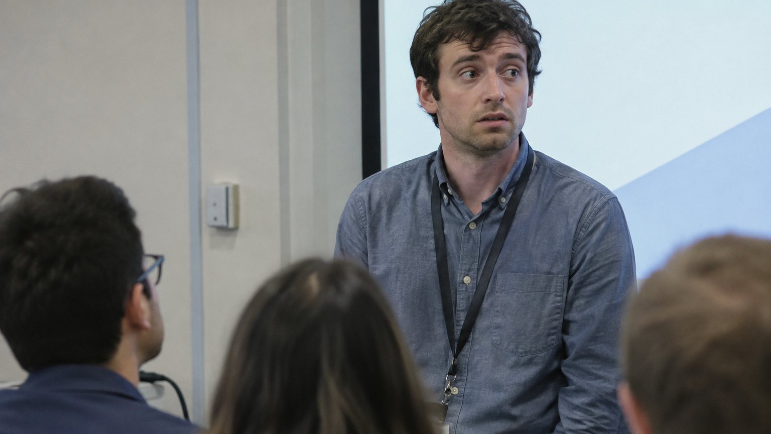A nervous presenter pauses beside a projected slide while several blurred audience members look toward him