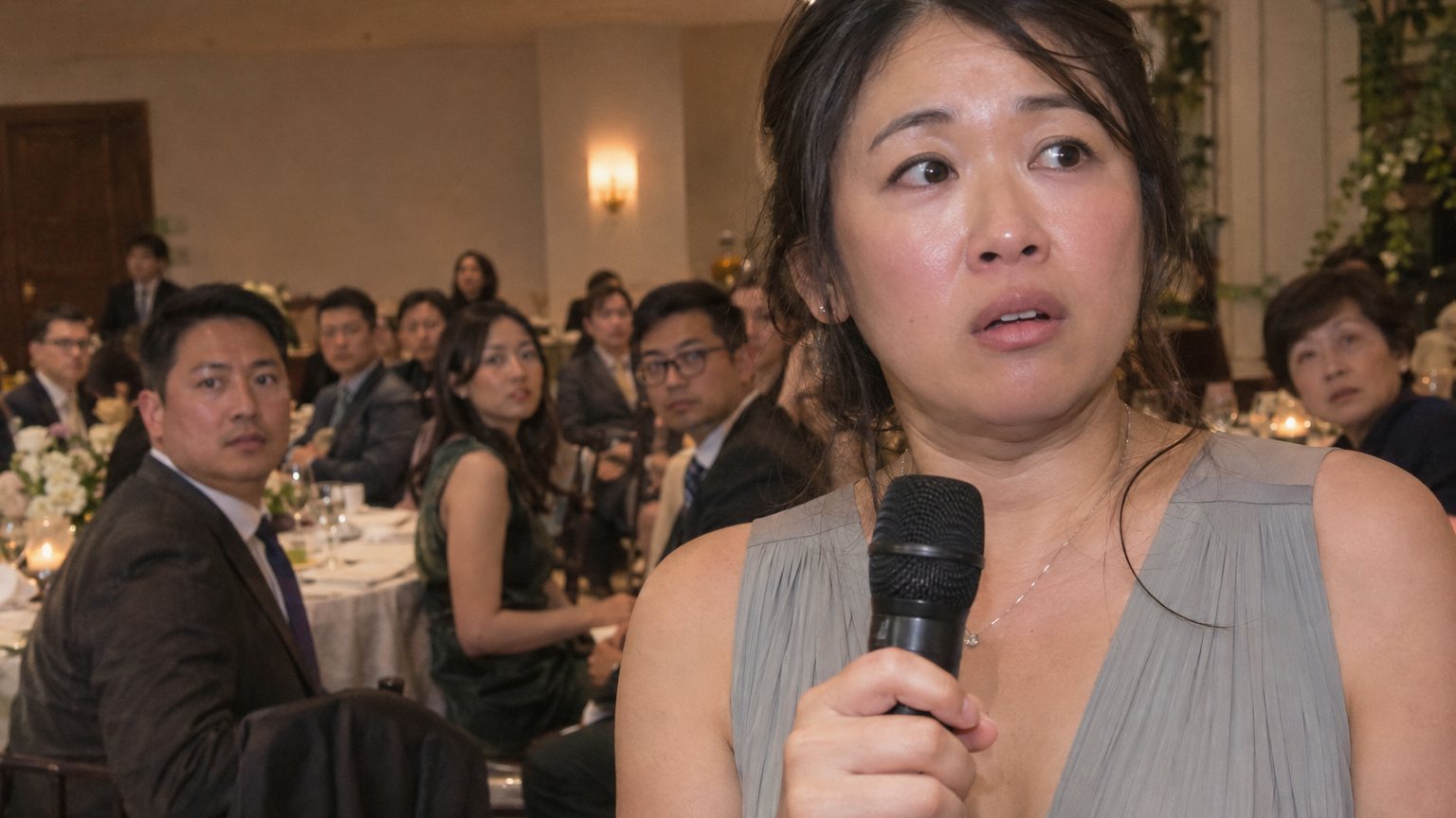 An East Asian woman freezes while holding a microphone during a wedding toast as guests turn toward her