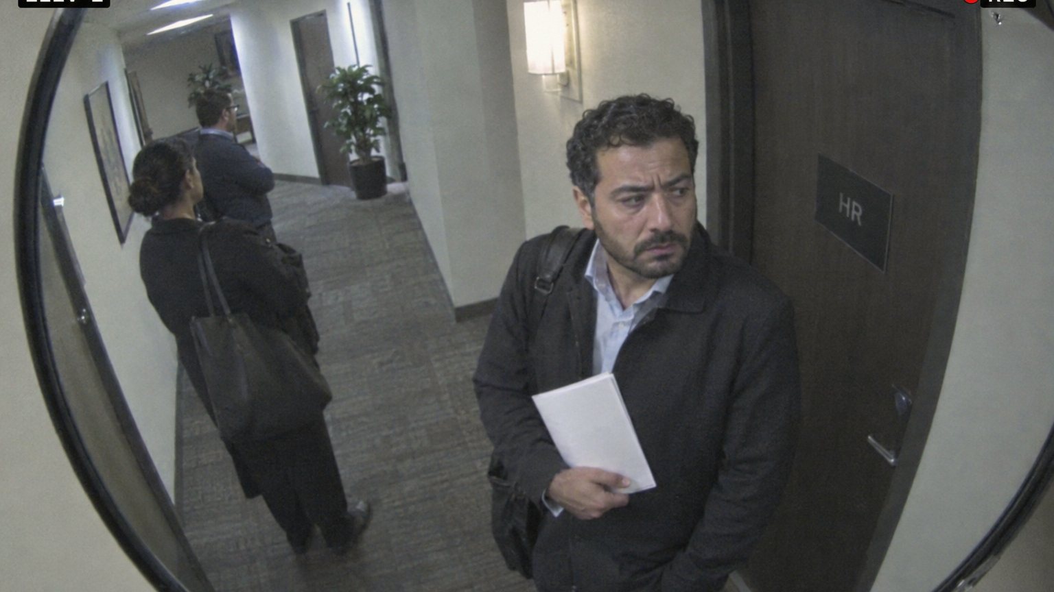 A Middle Eastern man reflected in a curved office hallway mirror waits outside HR holding a folded review document