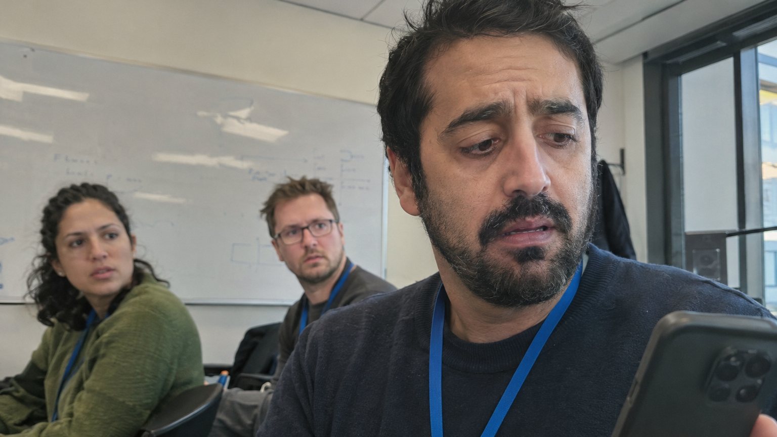 A man freezes while holding a ringing phone during a small team stand-up as colleagues wait by a whiteboard