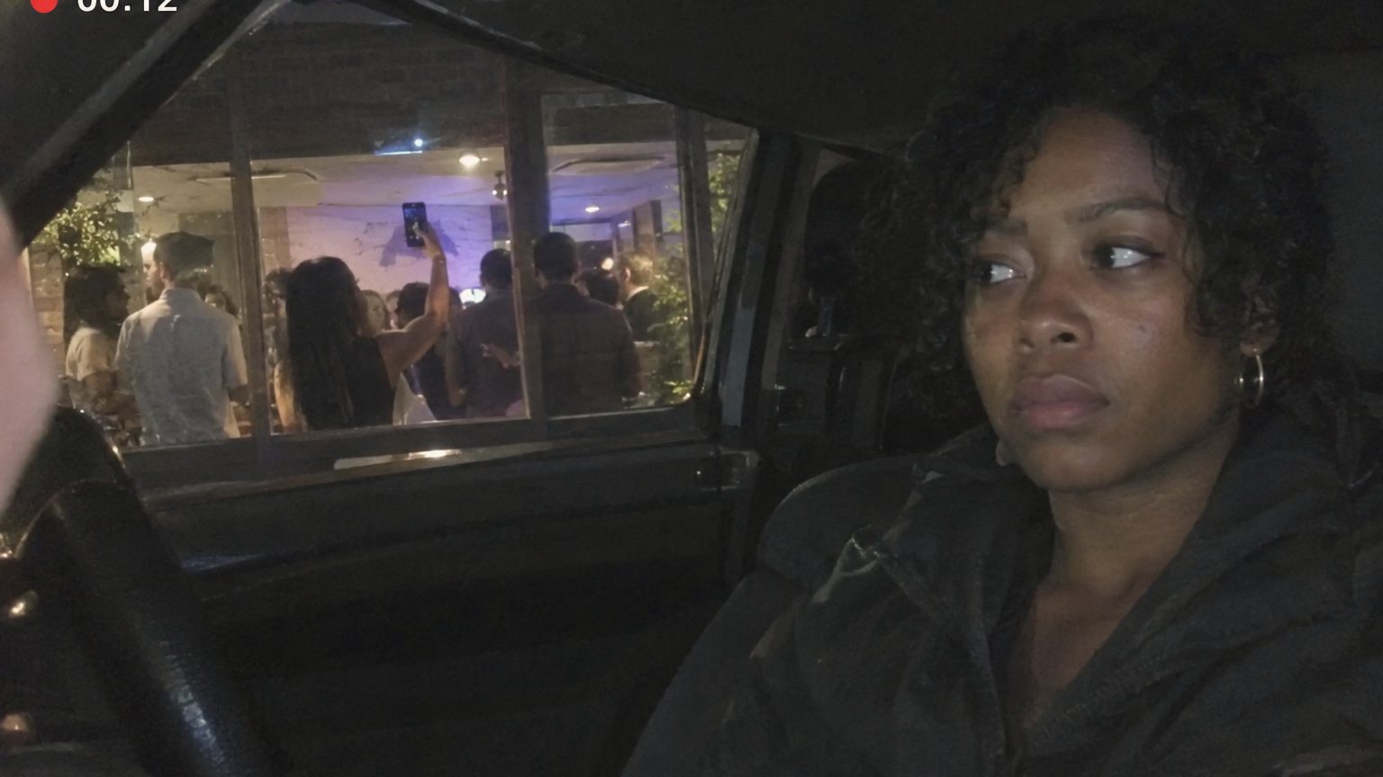 A Black woman sits tensely in a parked car outside a coworker party while a raised phone is visible inside the venue