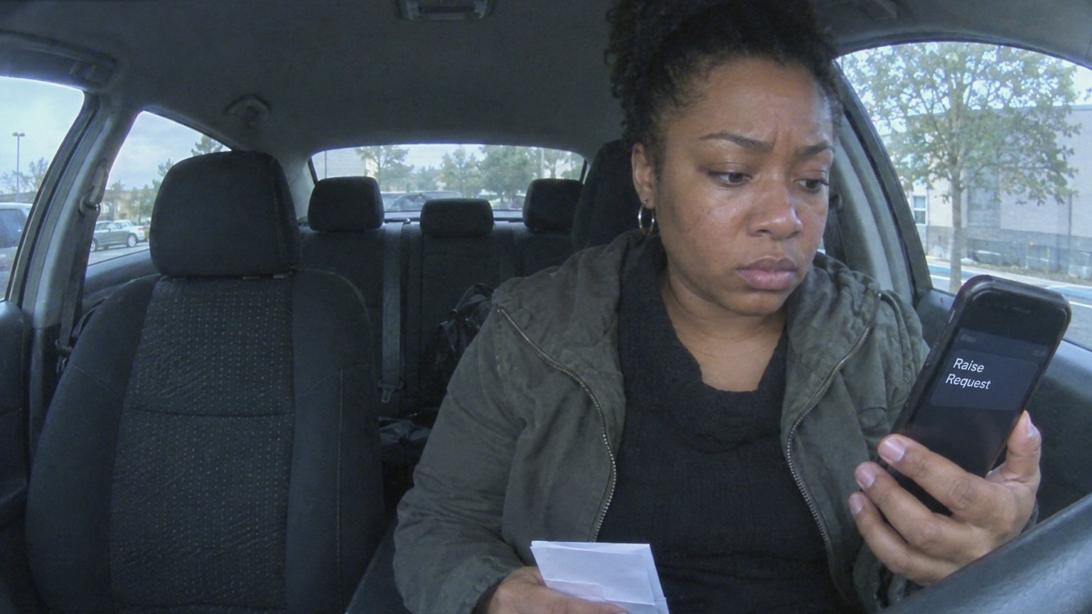A Black woman sits in a parked car before a pay conversation, holding a phone with an unreadable raise request